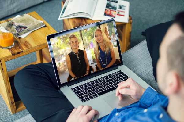 A person on a couch looks at a laptop screen displaying a video call with a smiling man and woman. magazines and a glass of orange juice are on the table beside them.