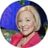 Profile picture of a smiling woman with short blonde hair, wearing a pink blazer, with a blurred nature background.