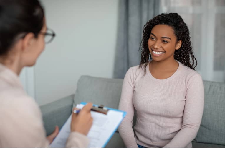 Two women sit on a couch; one is holding a clipboard and taking notes, the other is smiling, suggesting a conversation or counseling session.