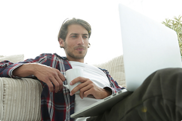 A man in a plaid shirt holds a coffee mug and uses a laptop while lounging on a sofa.