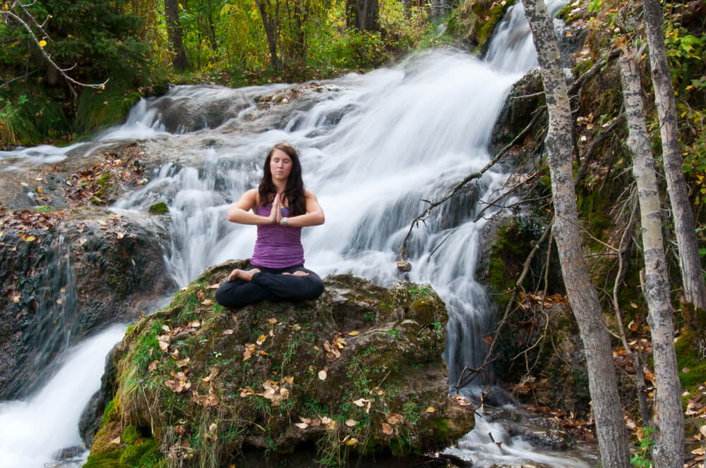 A woman meditating on a rock in front of a waterfall surrounded by forest.