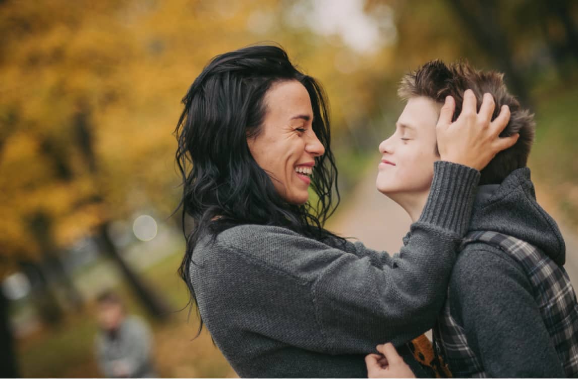 A woman smiles and touches a boy's head while they stand outdoors in a park with autumn foliage.