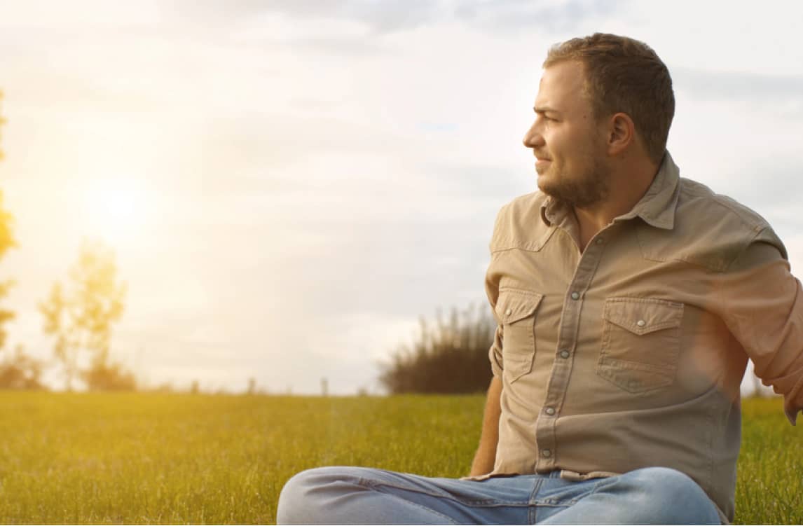 A man sits on grass outdoors, looking to the right as sunlight shines in the background. He is wearing a beige shirt and jeans.