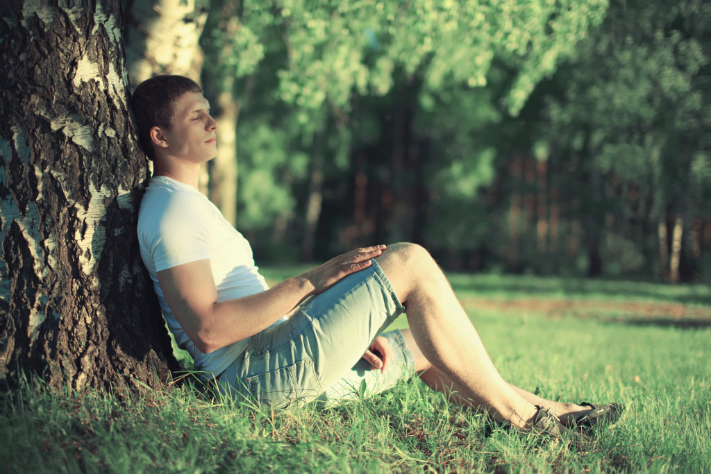 Pensive man sitting near a tree with his eyes closed meditating and enjoying the warm summer sunset