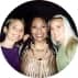 Three young women smiling closely together for a photo, one wearing purple, one in a beige top, and one in green.