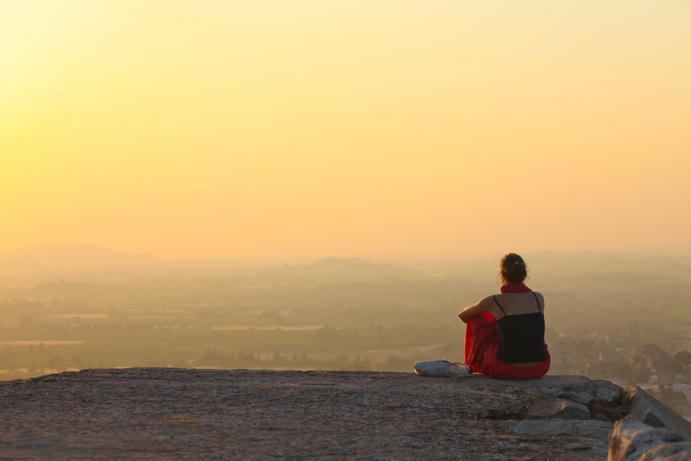Person sitting on a rock overlooking a scenic landscape at sunset.