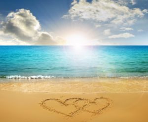 A heart drawn in the sand on a sunny beach with clear blue water and a sky with scattered clouds.