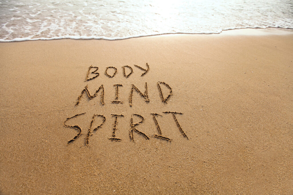 The words "Body Mind Spirit" are written in the sand near the edge of a wave on a beach.