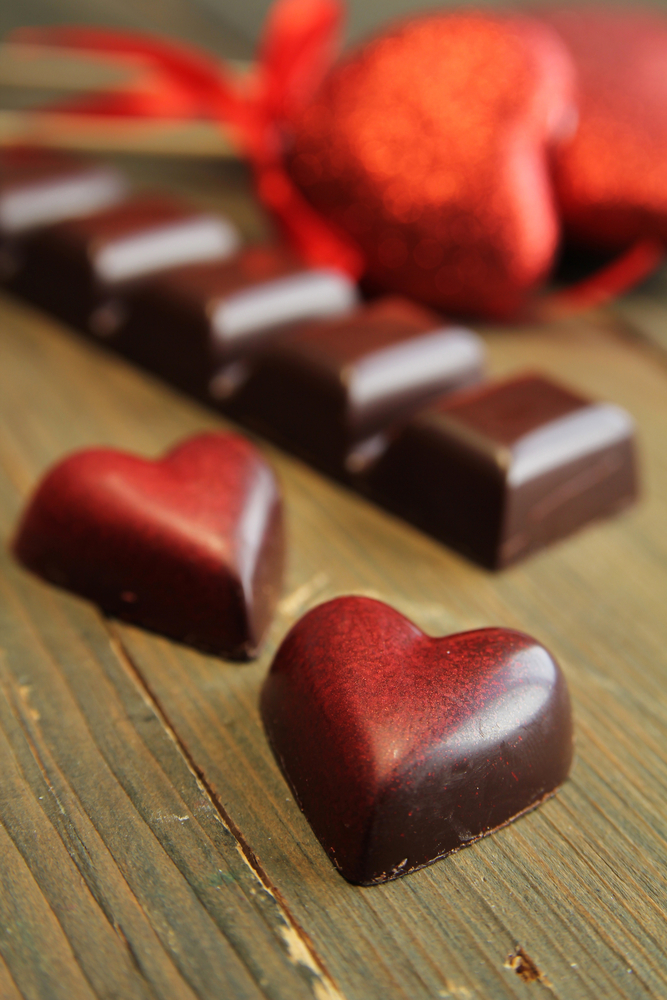 Rows of chocolate candies in the shape of hearts on a wooden surface, with a glittery red heart ornament in the background.