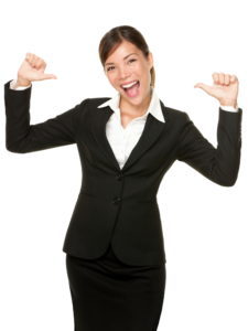 A smiling woman in business attire with thumbs pointing towards herself.