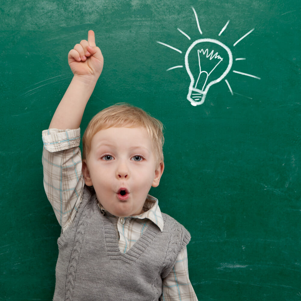 A young boy gestures having an idea in front of a chalkboard with a lightbulb drawn on it.