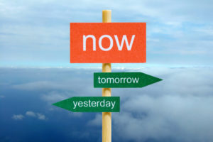 Signpost with the words "now," "tomorrow," and "yesterday" pointing in different directions against a cloudy sky background.