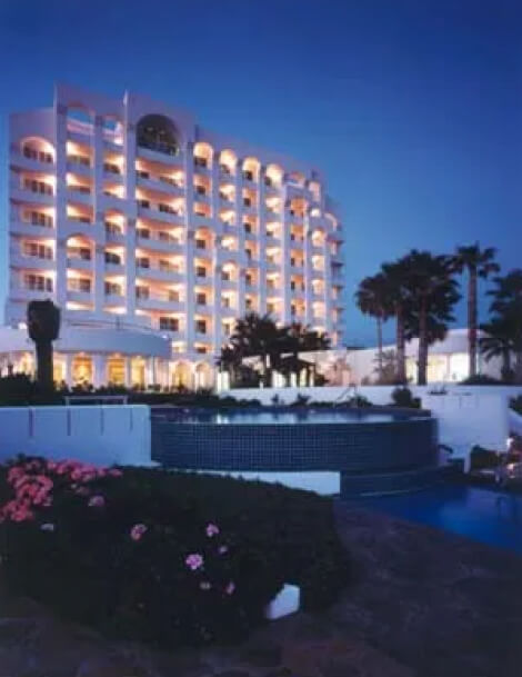 A multi-story hotel building with illuminated balconies at dusk, surrounded by palm trees, a pool, and landscaped gardens.