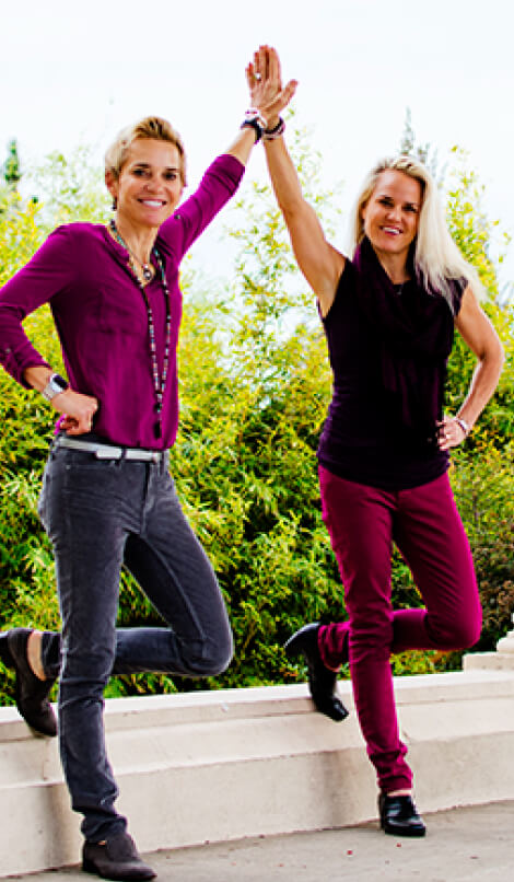 Two women stand on one leg while high-fiving outdoors. Both are smiling and wearing casual outfits in shades of purple. Green foliage is visible in the background.