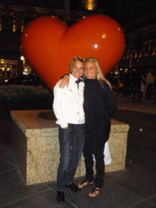 Two people smiling in front of a large heart-shaped sculpture at night.
