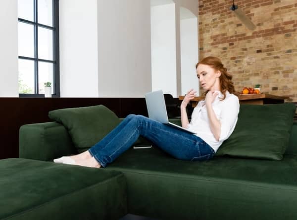 A woman in jeans and a white shirt sits on a green sofa with a laptop, looking thoughtful.