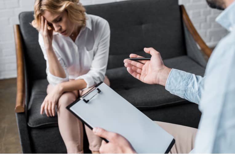 A woman sits on a couch with her head in her hand, appearing distressed, while a person with a clipboard speaks to her during a counseling session.