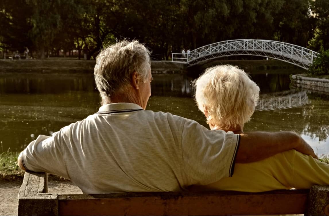 Two elderly people sit closely together on a bench by a pond, facing a small arched footbridge in a park setting.