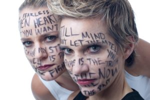Two women with words written on their faces looking intently at the camera.