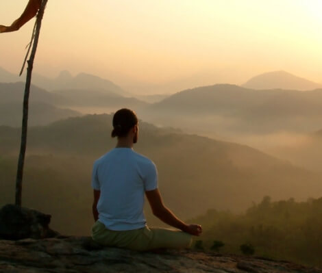 A person sits cross-legged on a rock, meditating at sunrise with misty hills and mountains in the background.