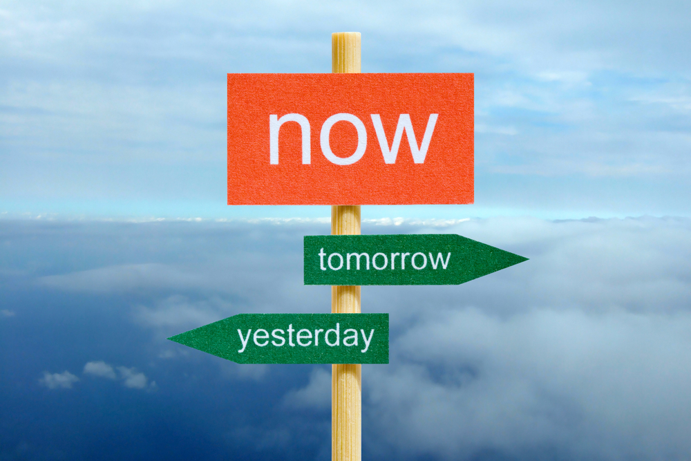 Signpost with the words "now," "tomorrow," and "yesterday" pointing in different directions against a cloudy sky background.
