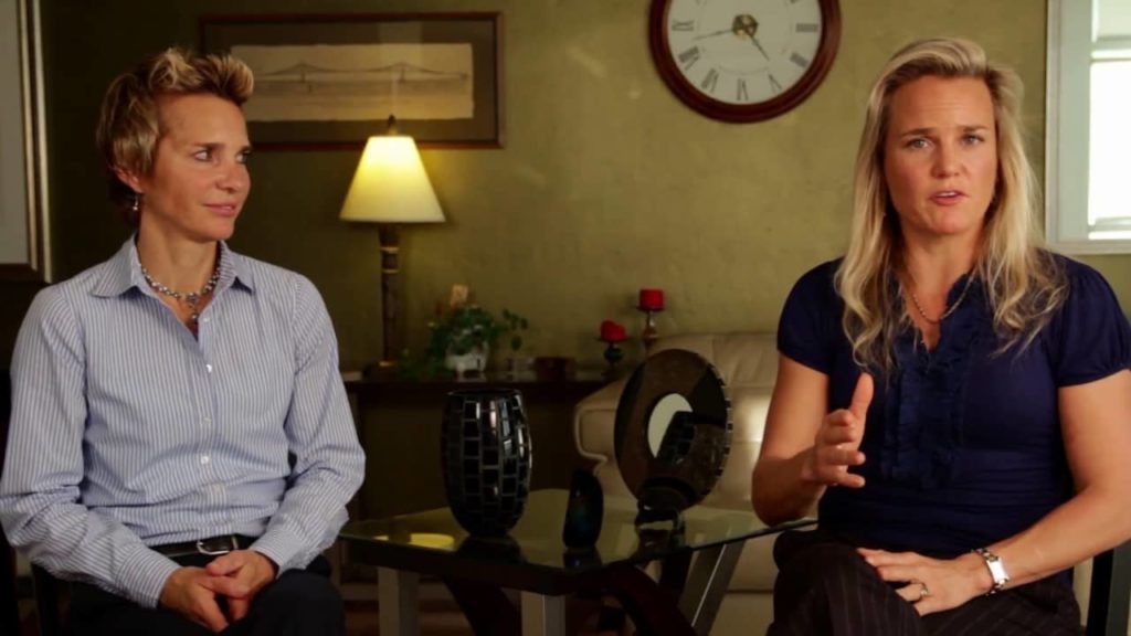 Two women seated with a table between them; one is speaking and gesturing, the other listens attentively. decor includes a lamp and a clock in the background.