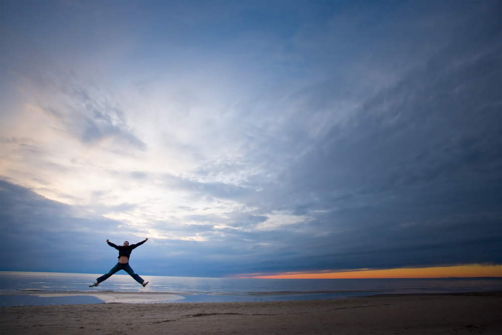 A person is captured mid-jump on a tranquil beach against a sunset sky.