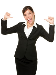 A smiling woman in business attire with thumbs pointing towards herself.