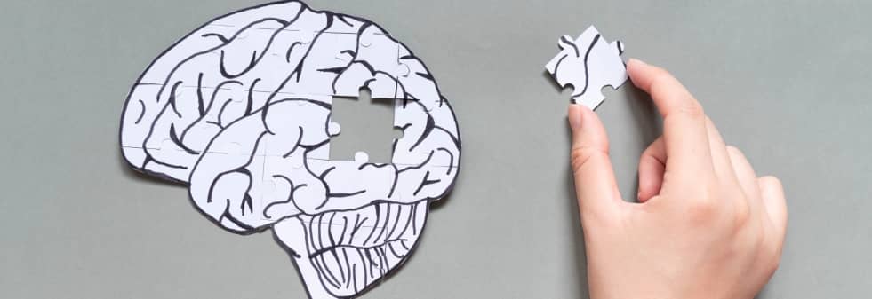 A hand placing the final piece into a jigsaw puzzle shaped like a human brain on a gray background.