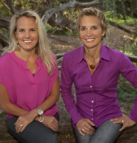Two women sit outdoors on tree branches. Both are smiling; one wears a pink blouse, the other a purple shirt with a heart necklace. The background features trees and greenery.