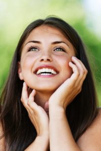 A woman smiling and looking upwards with her hands on her face against a blurred green background.