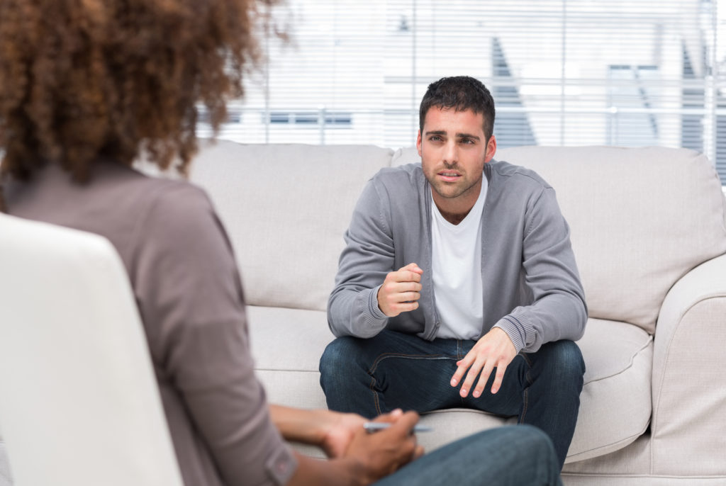A man seated on a sofa gestures while speaking to someone opposite him who is only partially visible in the foreground.