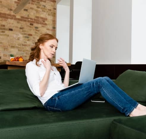 A woman sitting on a green sofa, using a laptop and holding a coffee mug in a modern apartment.