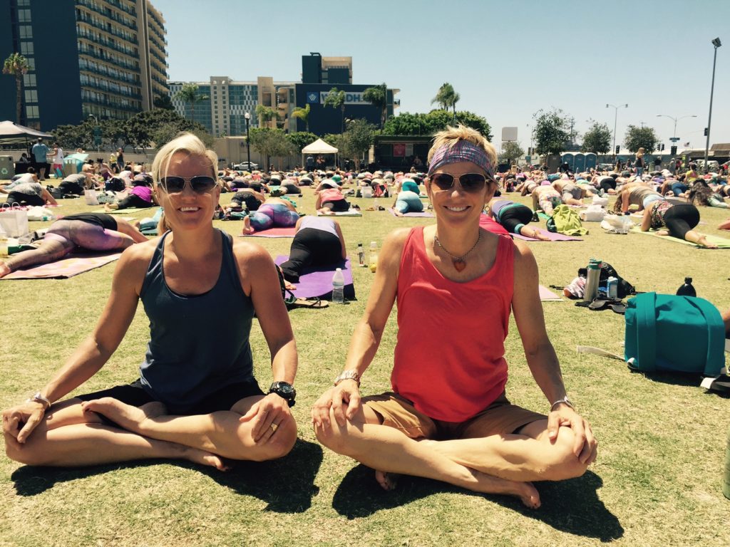 Two individuals seated cross-legged on the grass at an outdoor yoga event, surrounded by other participants in a sunny urban park setting.