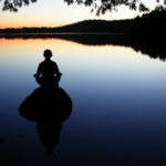 person meditating on a rock at dusk overlooking lake