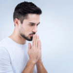Sad young man praying while standing against the blue background Sad young man praying while standing against the blue background