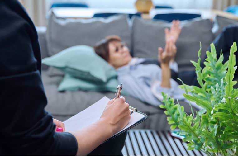 A person reclines on a couch talking while another person, holding a clipboard and pen, takes notes in the foreground. A green plant is visible on a table nearby.