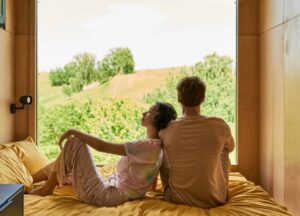 What Is Your Attachment Style? Understand How We Connect, Relate, and Heal Two people sit on a bed facing a large window, looking out at green trees and grassy hills under a bright sky.