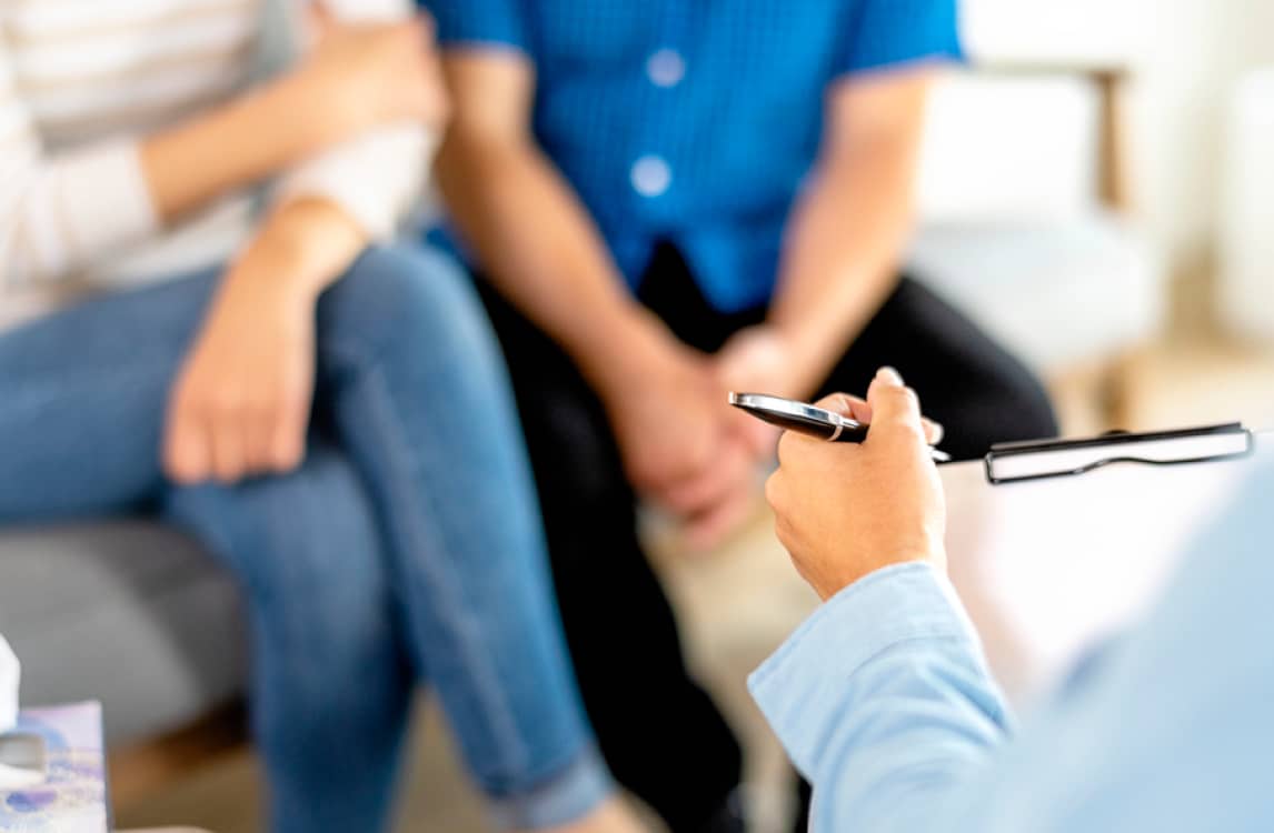 A therapist holding a pen speaks to a seated couple during a counseling session.