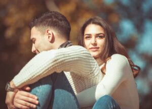 Healing Attachment Wounds with Brainspotting: How Compassion, Co-Regulation, and Subcortical Processing Rewire the Brain A woman with long brown hair hugs a man from behind as they sit outdoors in sunlight, both wearing white sweaters and blue jeans.