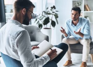 Trauma Informed Therapy Two men sit in an office; one takes notes in a notebook while the other gestures with his hands, suggesting a therapy or counseling session.