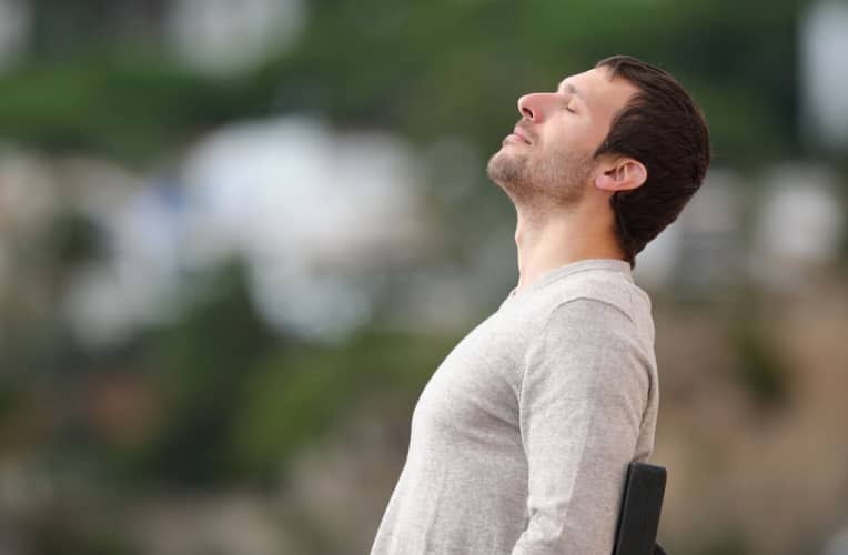 A man sits outdoors with his head tilted back and eyes closed, appearing relaxed, against a blurred green and white background.