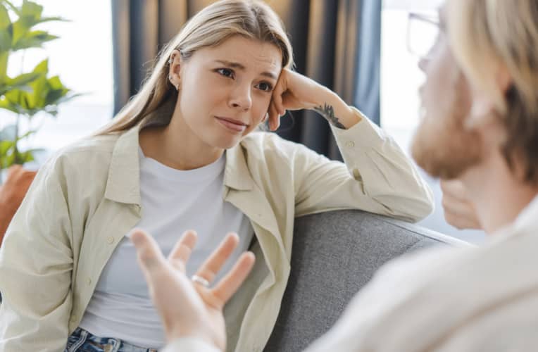 A woman sits on a couch listening intently to a person speaking, with her head resting on her hand and a concerned expression on her face.